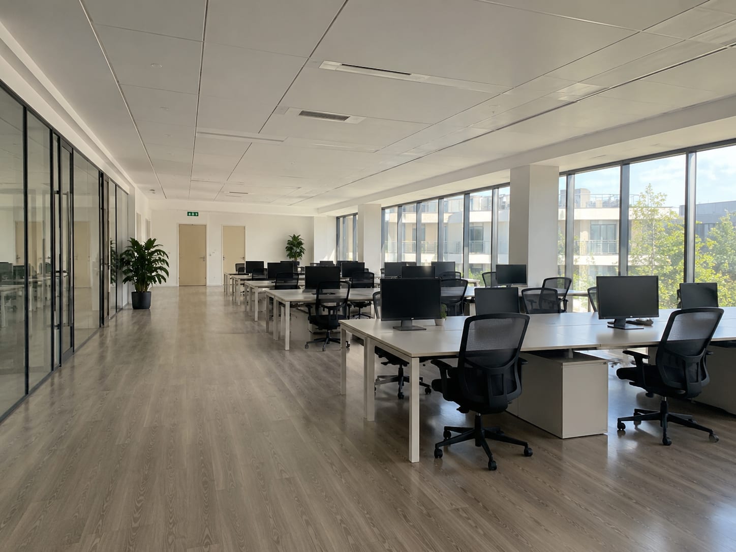 A large open-plan office after an out-of-hours clean: empty white bench desks with monitors, chairs tucked in, glass partition wall along one side, floor-to-ceiling windows and a wood floor with no marks on it.